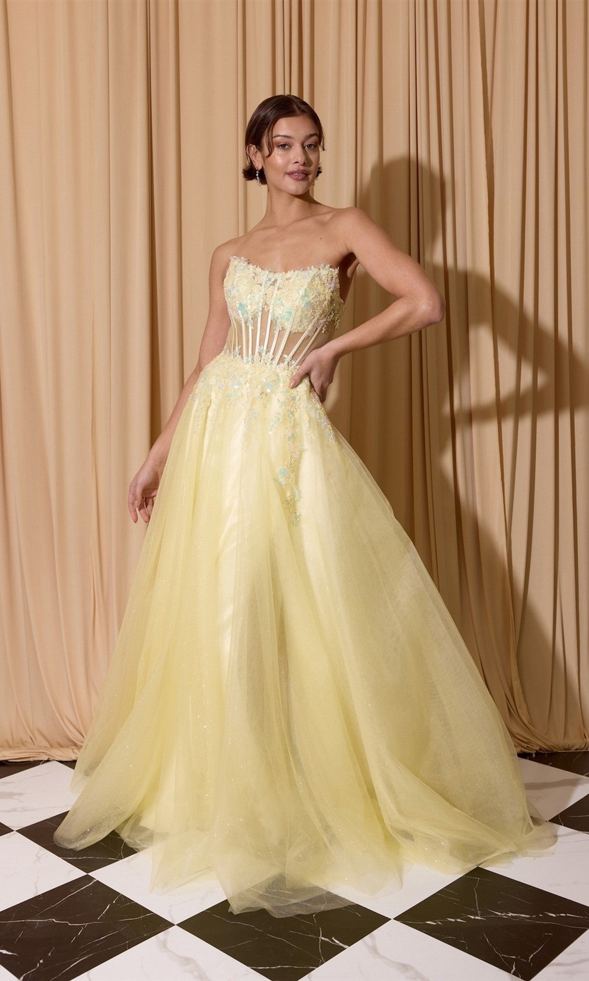 A woman stands against beige curtains wearing the Long Prom Dress: Eureka EK208—a floor-length yellow gown featuring a strapless sheer corset bodice, glitter tulle, and beaded floral appliqué embellishments.